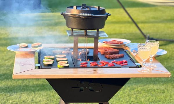 Zdjęcie przedstawia grill. Widoczne jest logo. Na grillu widoczne jest przygotowywane jedzenie, w tym warzywa oraz mięso. Na drewnianym dodatku do grilla postawione są talerze i kryształy z napojem w środku.