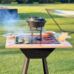 Zdjęcie przedstawia grill. Widoczne jest logo. Na grillu widoczne jest przygotowywane jedzenie, w tym warzywa oraz mięso. Na drewnianym dodatku do grilla postawione są talerze i kryształy z napojem w środku.