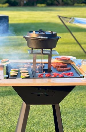 Zdjęcie przedstawia grill. Widoczne jest logo. Na grillu widoczne jest przygotowywane jedzenie, w tym warzywa oraz mięso. Na drewnianym dodatku do grilla postawione są talerze i kryształy z napojem w środku.