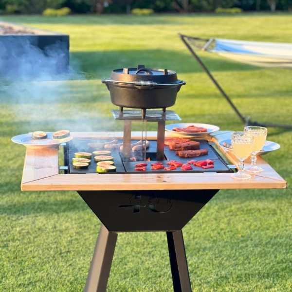 Zdjęcie przedstawia grill. Widoczne jest logo. Na grillu widoczne jest przygotowywane jedzenie, w tym warzywa oraz mięso. Na drewnianym dodatku do grilla postawione są talerze i kryształy z napojem w środku.